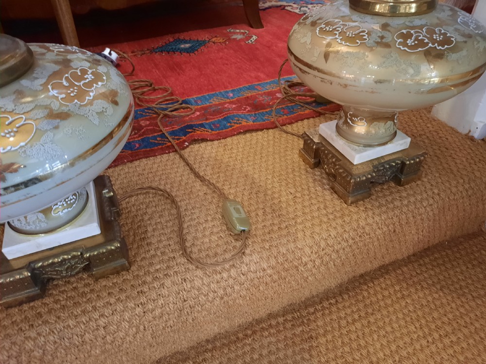 A pair of late Victorian table lamps featuring ornate gilt brass stems, painted glass baluster bases, and cream silk shades with fringe tassels. View VI