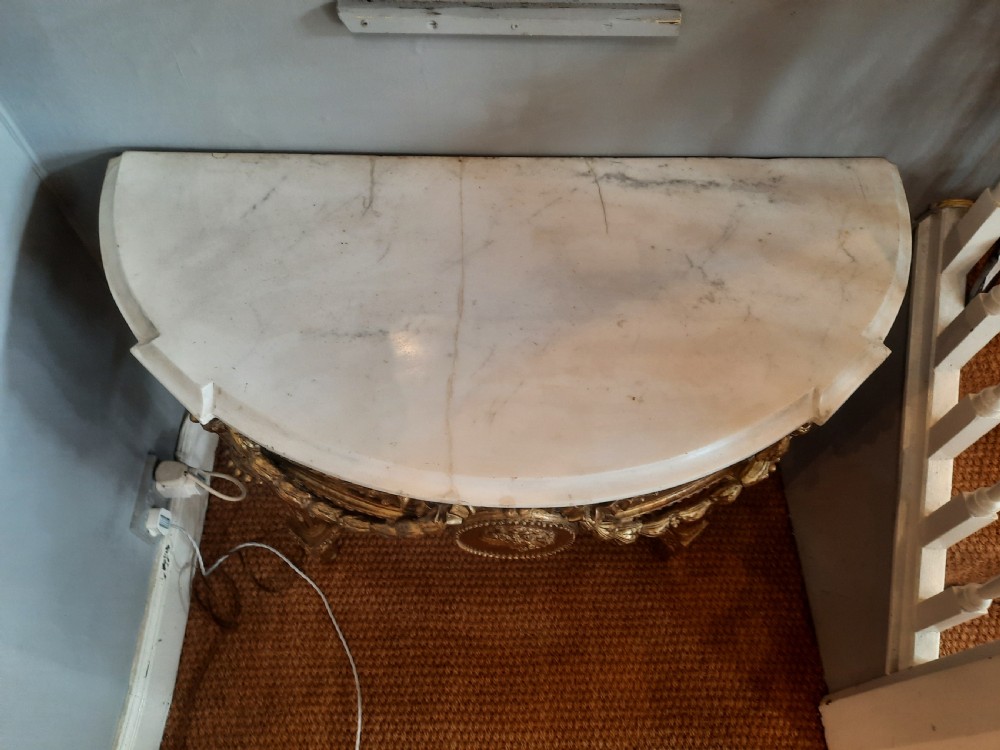 An early 19th-century semicircular giltwood console table featuring a white marble top, carved classical medallion, decorative swags, and fluted legs. View VII