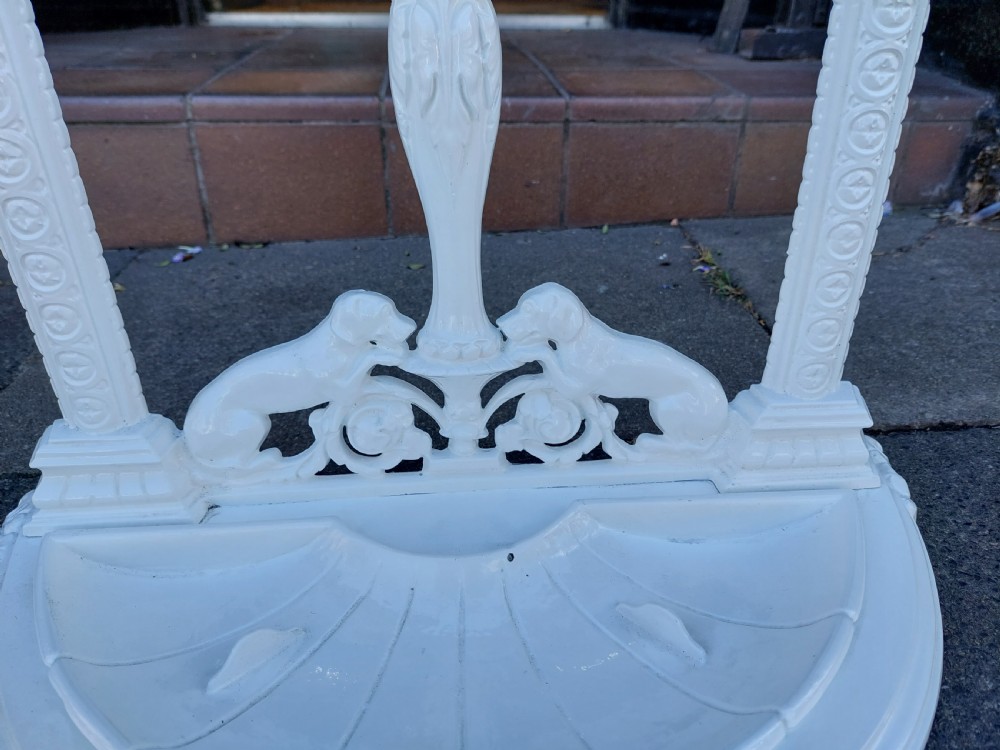 A white painted cast iron Victorian stick stand featuring detailed scrollwork, an arched backplate frame with a top ring, a central pedestal, and a curved drip tray base. View III