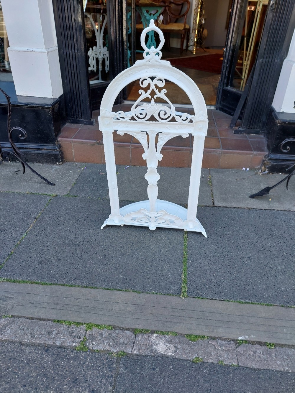 A white painted cast iron Victorian stick stand featuring detailed scrollwork, an arched backplate frame with a top ring, a central pedestal, and a curved drip tray base. View VII