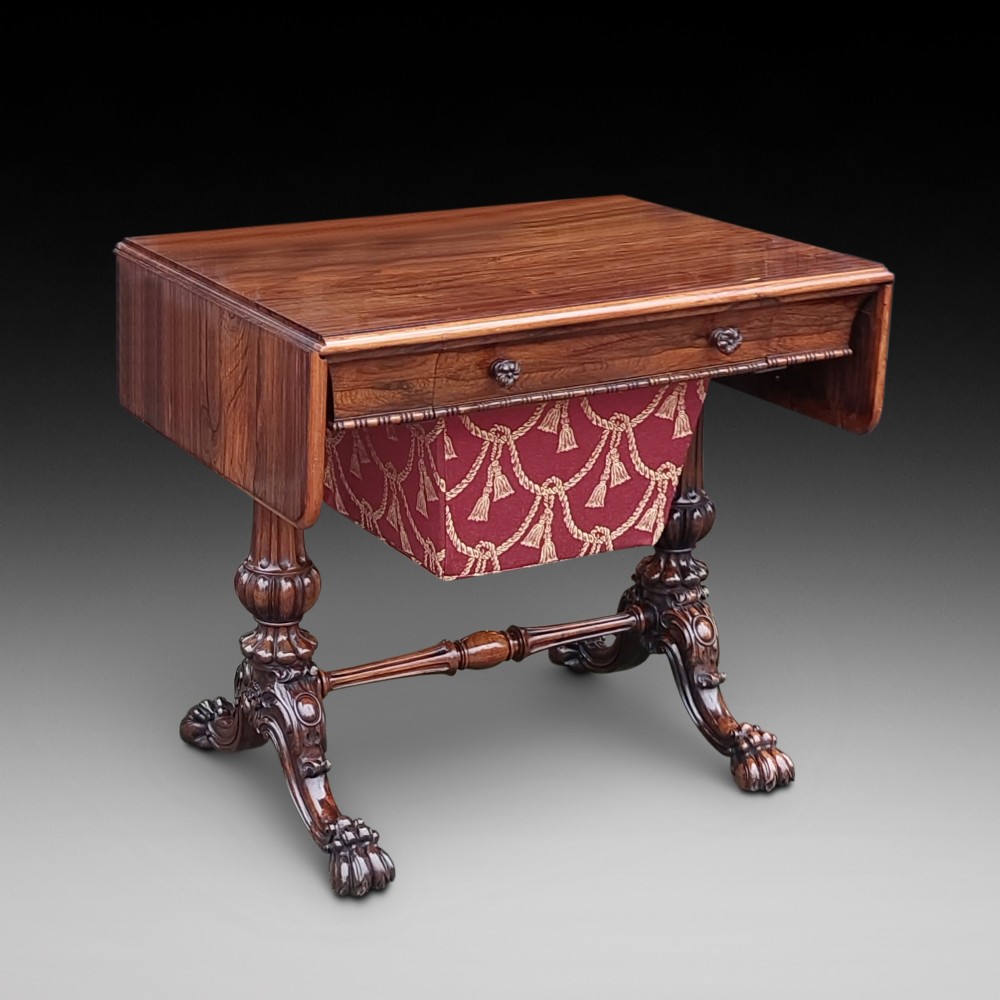 A Regency rosewood work table featuring drop leaves, a single frieze drawer, a red patterned fabric sewing well below, turned pedestal supports, and carved claw feet.