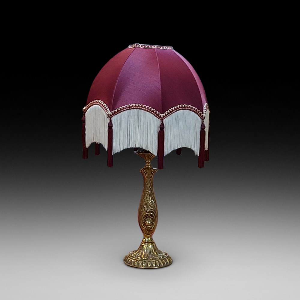An early 20th-century table lamp featuring an embossed pressed brass stem and a dome-shaped red fabric shade trimmed with cream fringe and tassels.
