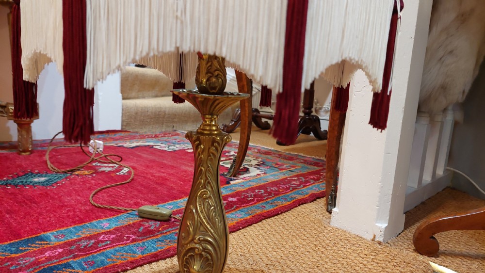 An early 20th-century table lamp featuring an embossed pressed brass stem and a dome-shaped red fabric shade trimmed with cream fringe and tassels. View V