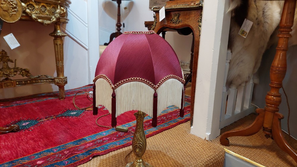 An early 20th-century table lamp featuring an embossed pressed brass stem and a dome-shaped red fabric shade trimmed with cream fringe and tassels. View VI