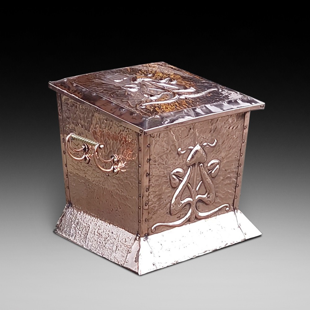 A square hand-hammered copper coal box featuring embossed stylised botanical motifs, riveted metal borders, side handles, and a flared base.