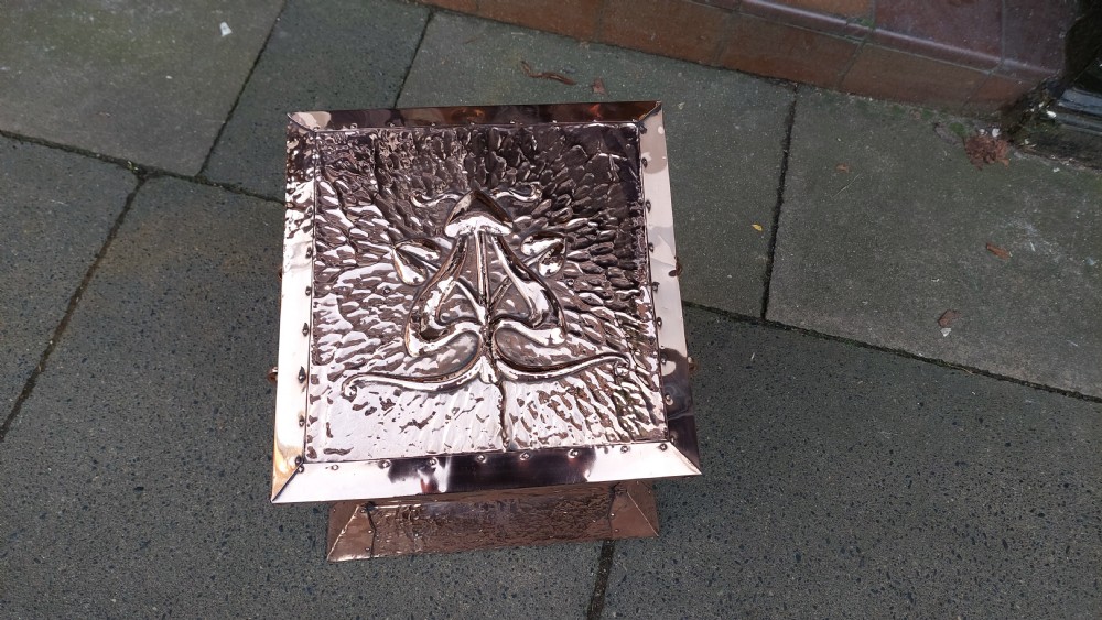 A square hand-hammered copper coal box featuring embossed stylised botanical motifs, riveted metal borders, side handles, and a flared base. View II