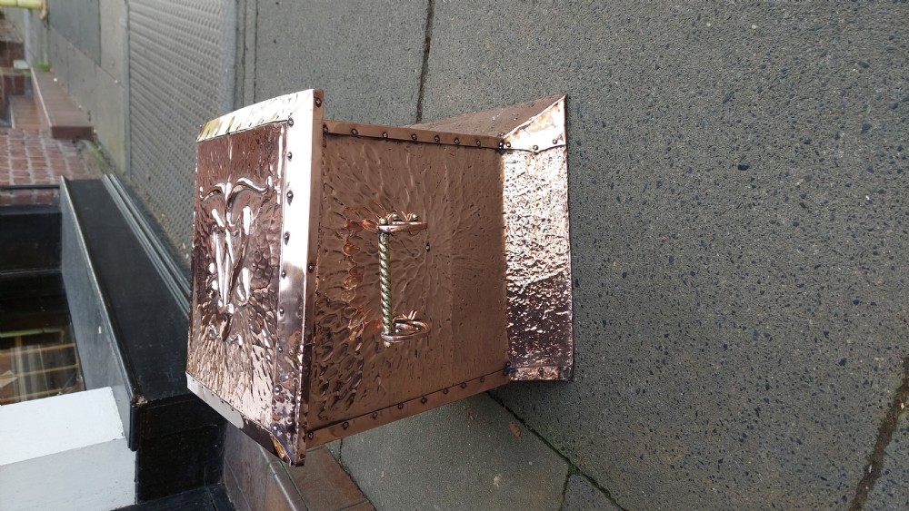 A square hand-hammered copper coal box featuring embossed stylised botanical motifs, riveted metal borders, side handles, and a flared base. View III