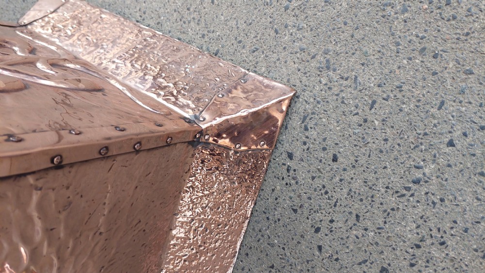 A square hand-hammered copper coal box featuring embossed stylised botanical motifs, riveted metal borders, side handles, and a flared base. View V