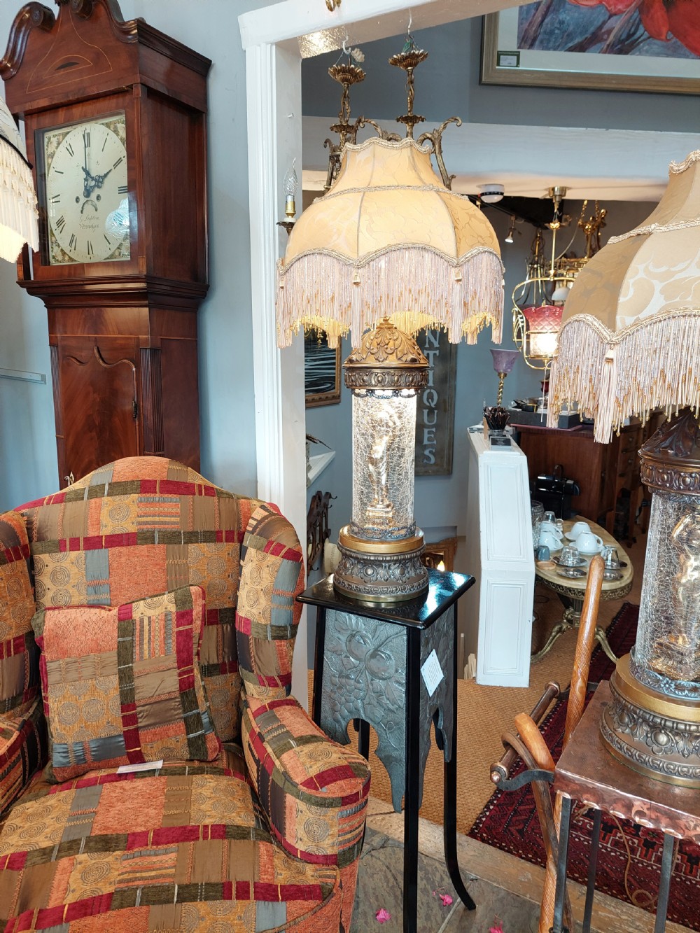 A pair of ornate gilded spelter table lamps featuring classical figures enclosed in glass columns topped with fringed cream fabric shades. View II