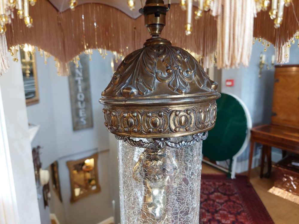 A pair of ornate gilded spelter table lamps featuring classical figures enclosed in glass columns topped with fringed cream fabric shades. View IV