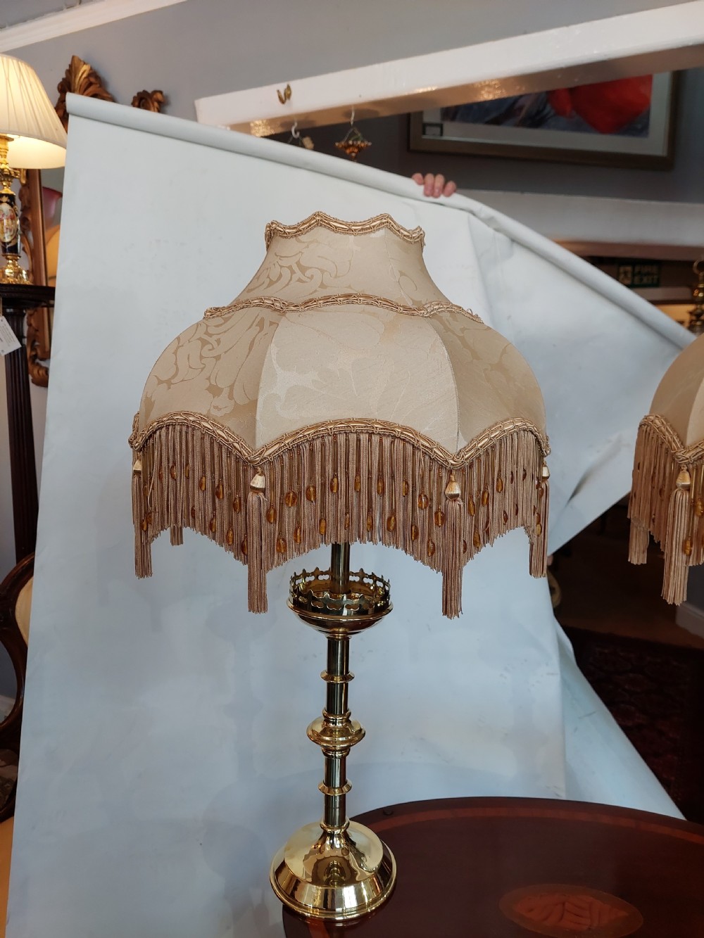 A pair of 19th-century Gothic Revival brass altar candlesticks converted into table lamps with patterned cream fabric shades featuring tassel fringe. View III