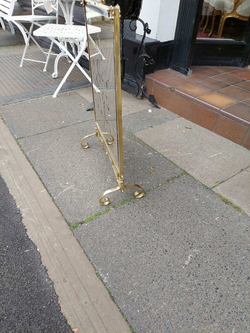 An Art Nouveau brass fire screen featuring a bevelled glass panel etched with a stylised floral motif, resting on splayed brass feet with a decorative top handle. View II