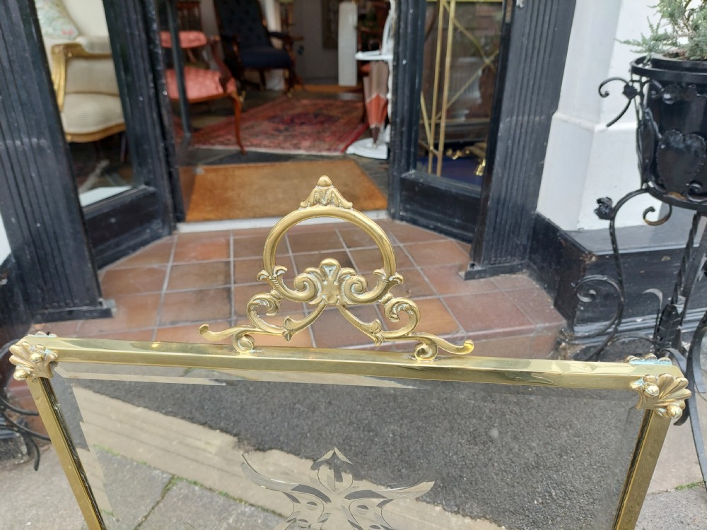 An Art Nouveau brass fire screen featuring a bevelled glass panel etched with a stylised floral motif, resting on splayed brass feet with a decorative top handle. View IV