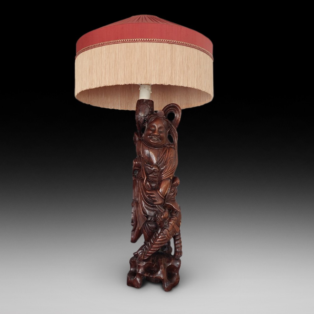An early 20th-century Chinese carved rootwood table lamp featuring a figural base and a cylindrical shade with cream fringe and red trim.