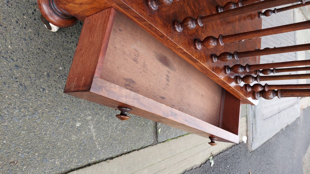 A Victorian walnut Canterbury featuring multiple open compartments formed by turned spindles, sitting above a single drawer with wooden knobs and turned legs on castors. View VI