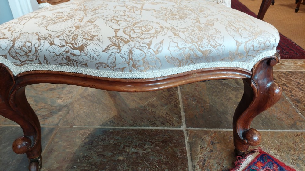 A Victorian low-seated nursing chair crafted from carved walnut featuring floral patterned cream fabric, spoon back, cabriole legs, and castors. View III
