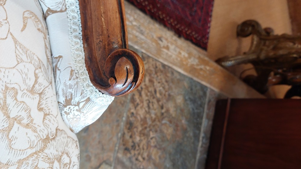A Victorian low-seated nursing chair crafted from carved walnut featuring floral patterned cream fabric, spoon back, cabriole legs, and castors. View V