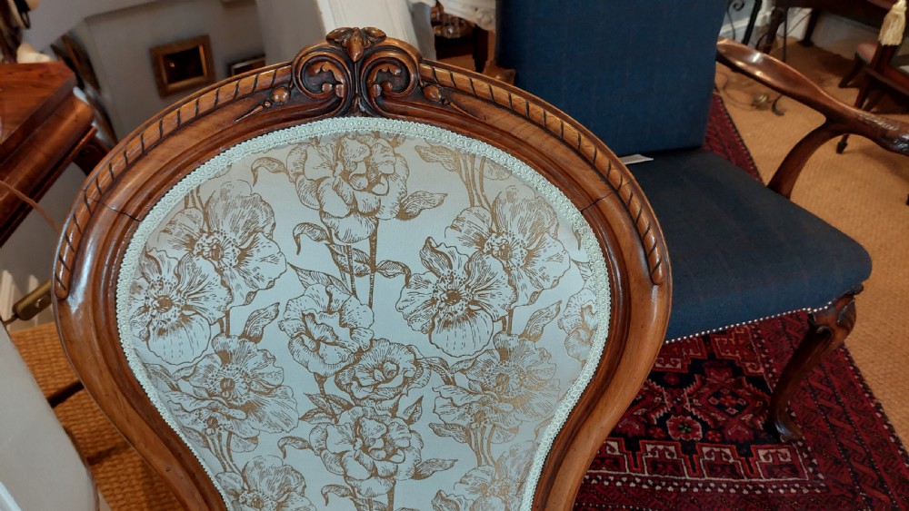 A Victorian low-seated nursing chair crafted from carved walnut featuring floral patterned cream fabric, spoon back, cabriole legs, and castors. View VI