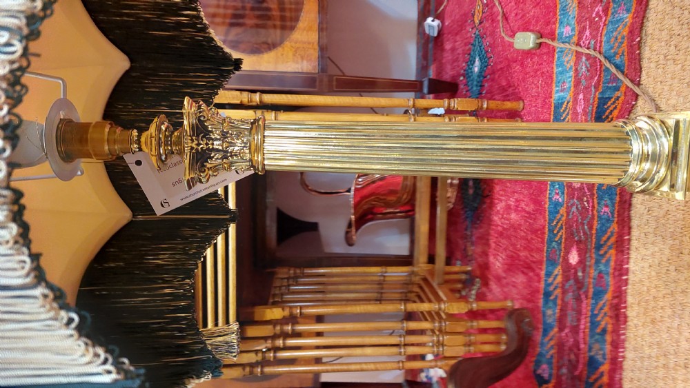 An early 20th-century brass table lamp featuring a fluted Corinthian column base on a stepped pedestal, topped with a cream domed shade trimmed with fringe and tassels. View IV