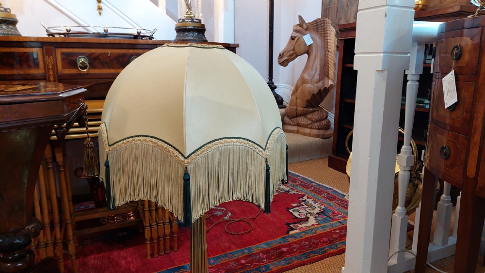 An early 20th-century brass table lamp featuring a fluted Corinthian column base on a stepped pedestal, topped with a cream domed shade trimmed with fringe and tassels. View V