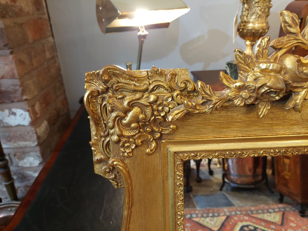 An upright rectangular wall mirror featuring an ornate gold gilt frame topped with a carved rocaille cresting motif and scrollwork corner details. View II