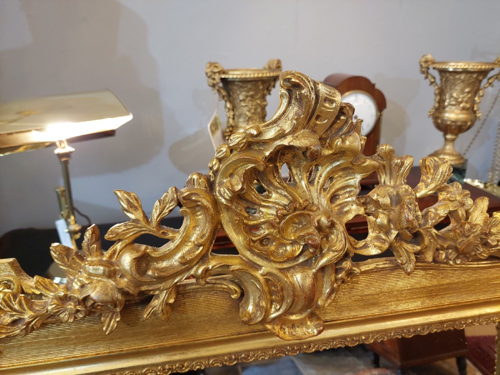 An upright rectangular wall mirror featuring an ornate gold gilt frame topped with a carved rocaille cresting motif and scrollwork corner details. View III