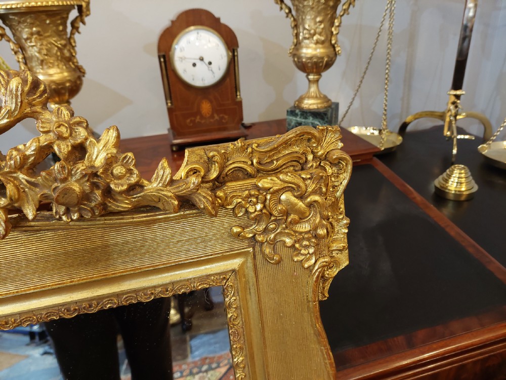 An upright rectangular wall mirror featuring an ornate gold gilt frame topped with a carved rocaille cresting motif and scrollwork corner details. View IV