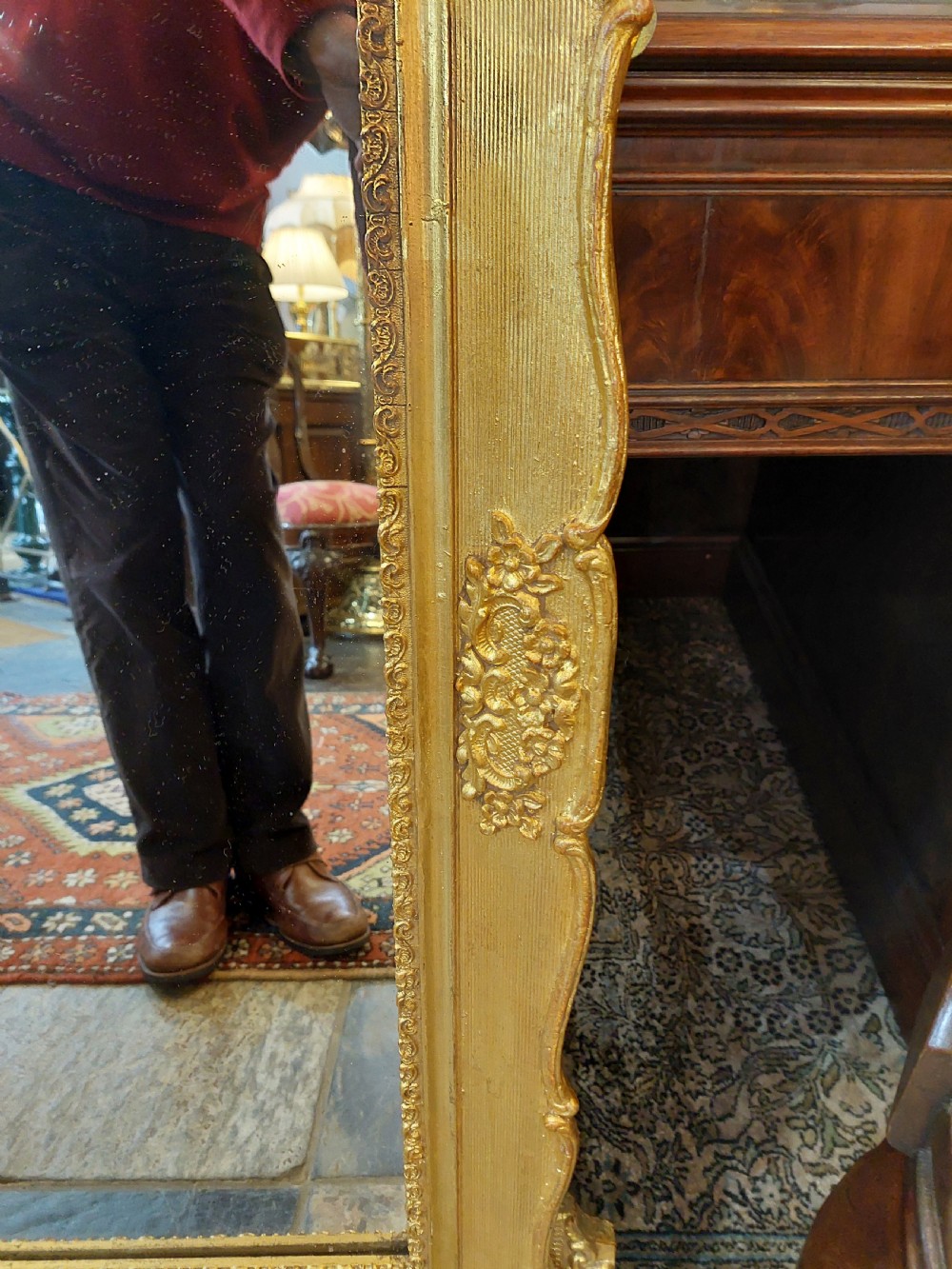 An upright rectangular wall mirror featuring an ornate gold gilt frame topped with a carved rocaille cresting motif and scrollwork corner details. View V