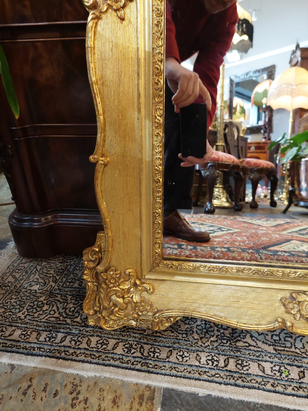An upright rectangular wall mirror featuring an ornate gold gilt frame topped with a carved rocaille cresting motif and scrollwork corner details. View VI