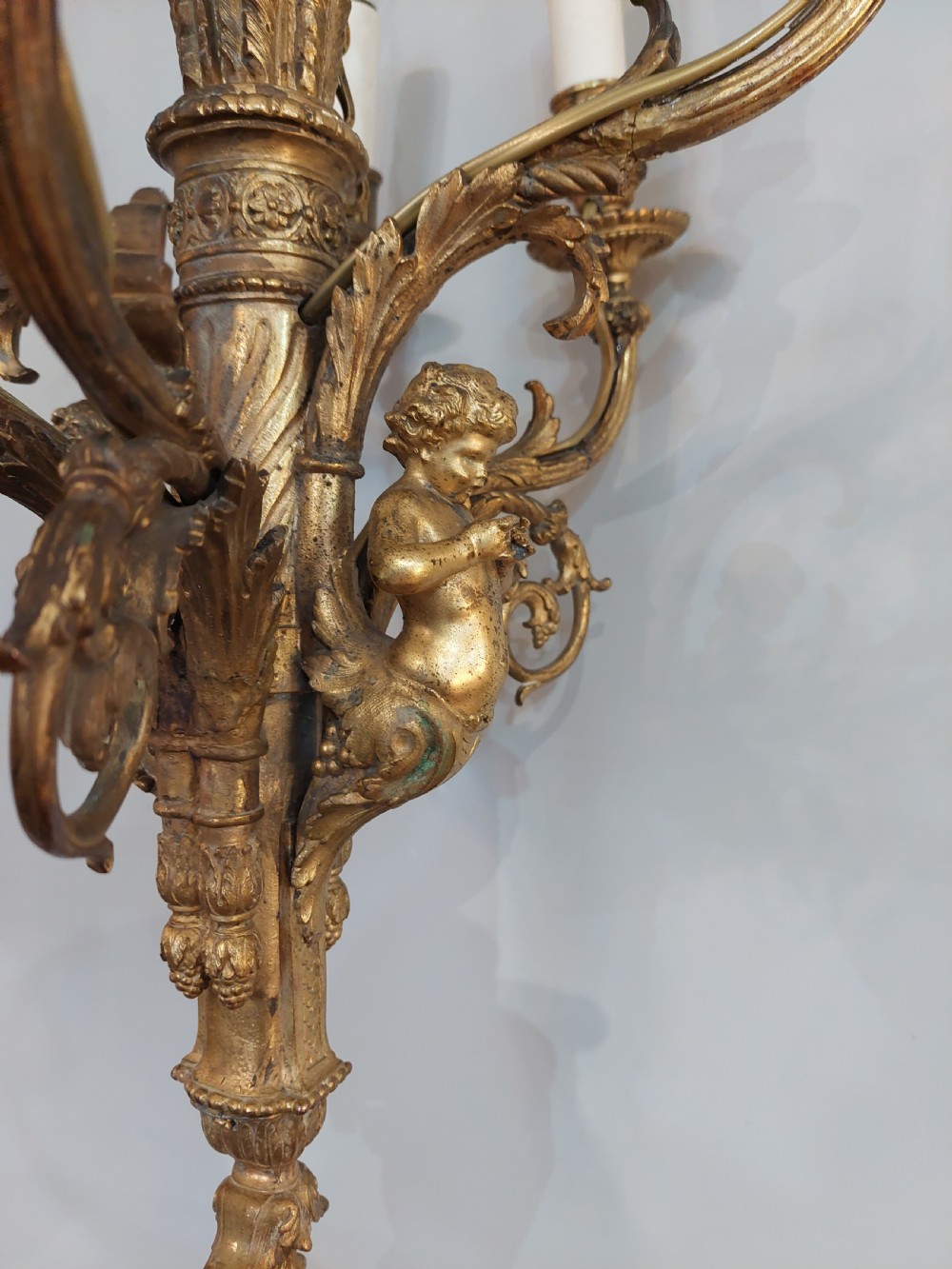 An early 20th-century six-arm gilt metal chandelier featuring carved cherub figures, scrolling acanthus leaf branches, and white candle sleeves. View IV