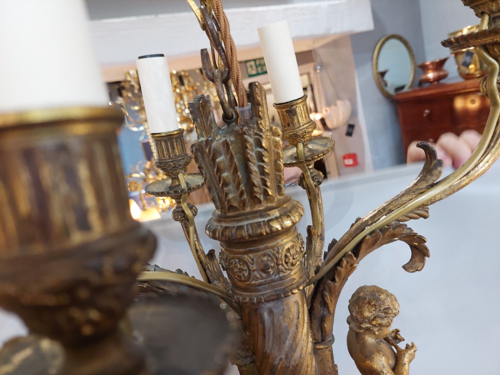 An early 20th-century six-arm gilt metal chandelier featuring carved cherub figures, scrolling acanthus leaf branches, and white candle sleeves. View X