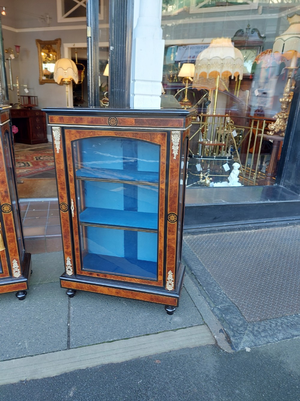 A pair of Victorian burr walnut display cabinets featuring ebonised borders, marquetry inlay, brass mounts, single glazed doors, and blue velvet lined shelves. View II