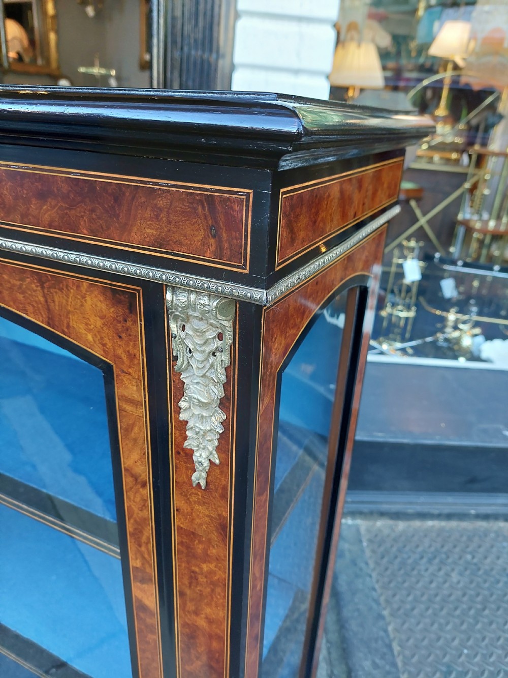 A pair of Victorian burr walnut display cabinets featuring ebonised borders, marquetry inlay, brass mounts, single glazed doors, and blue velvet lined shelves. View IV