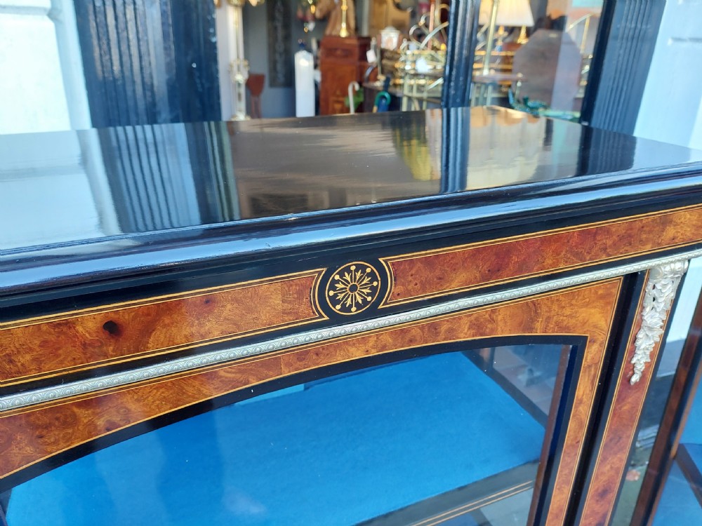 A pair of Victorian burr walnut display cabinets featuring ebonised borders, marquetry inlay, brass mounts, single glazed doors, and blue velvet lined shelves. View V