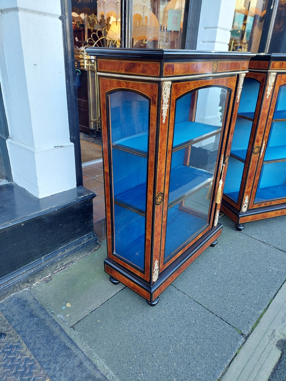 A pair of Victorian burr walnut display cabinets featuring ebonised borders, marquetry inlay, brass mounts, single glazed doors, and blue velvet lined shelves. View VI