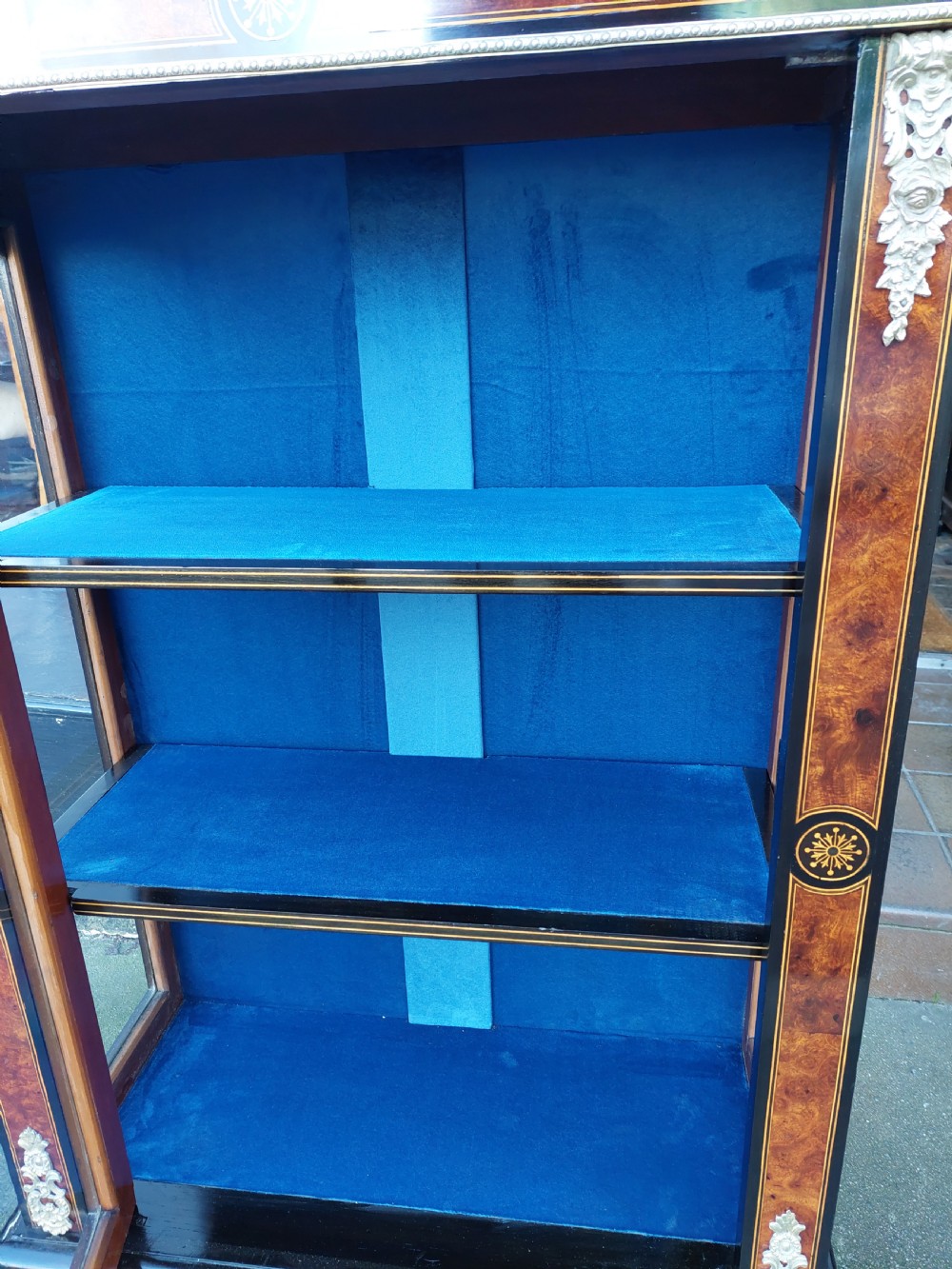 A pair of Victorian burr walnut display cabinets featuring ebonised borders, marquetry inlay, brass mounts, single glazed doors, and blue velvet lined shelves. View VIII