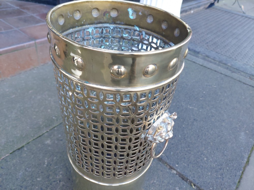 A cylindrical Victorian brass stick stand featuring a pierced lattice midsection, riveted top rim, and a central lion mask ring handle. View XII