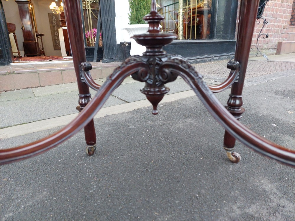 A late 19th-century mahogany centre table featuring a shaped top with marquetry inlay, turned legs, crossed stretcher with central finial, and brass castors. View V