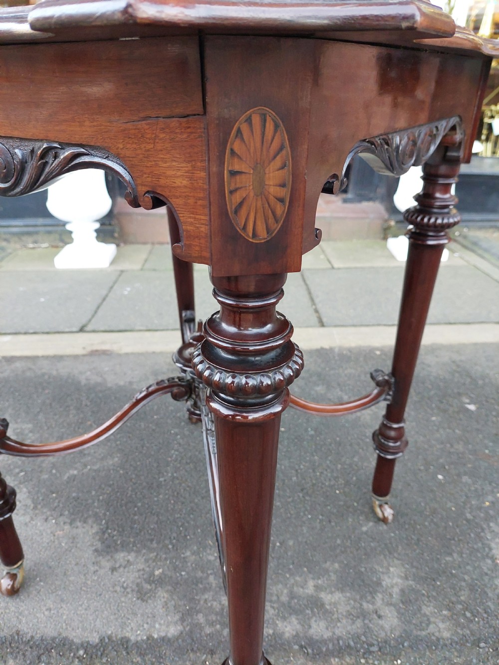 A late 19th-century mahogany centre table featuring a shaped top with marquetry inlay, turned legs, crossed stretcher with central finial, and brass castors. View VI