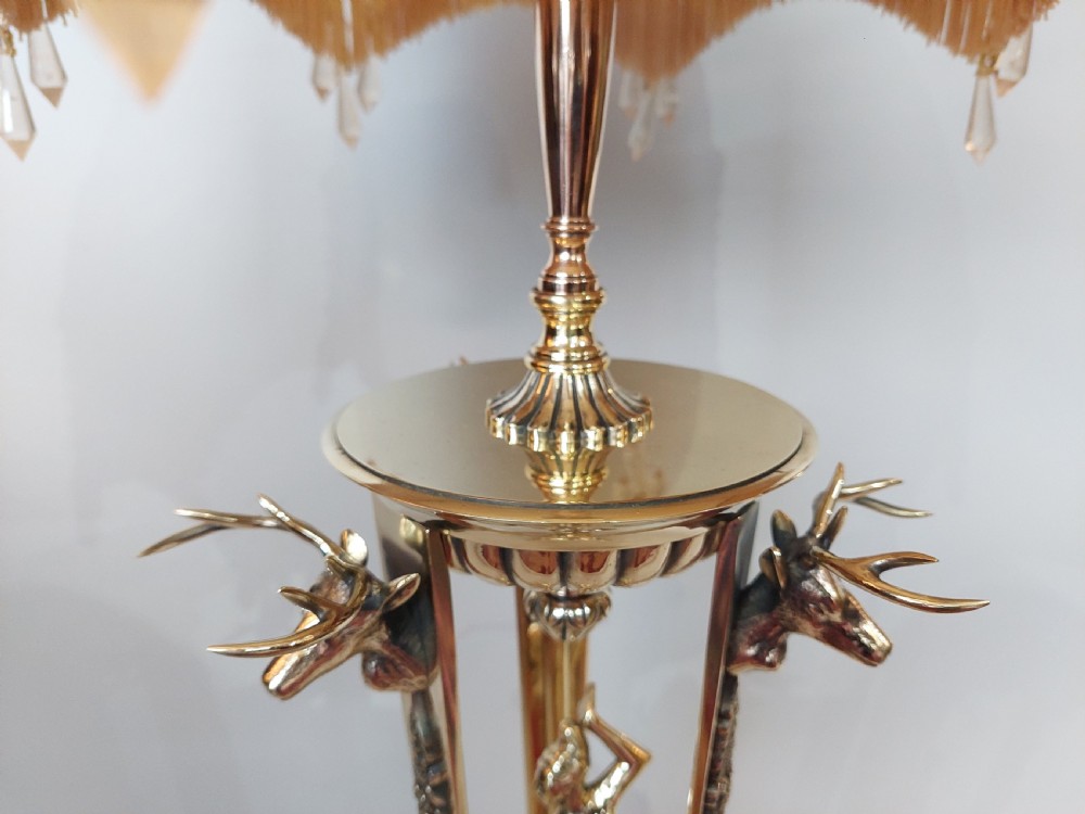 An early 20th-century brass table lamp featuring a central figure of Minerva flanked by stag head mounts, topped with a cream fringed lampshade. View VI