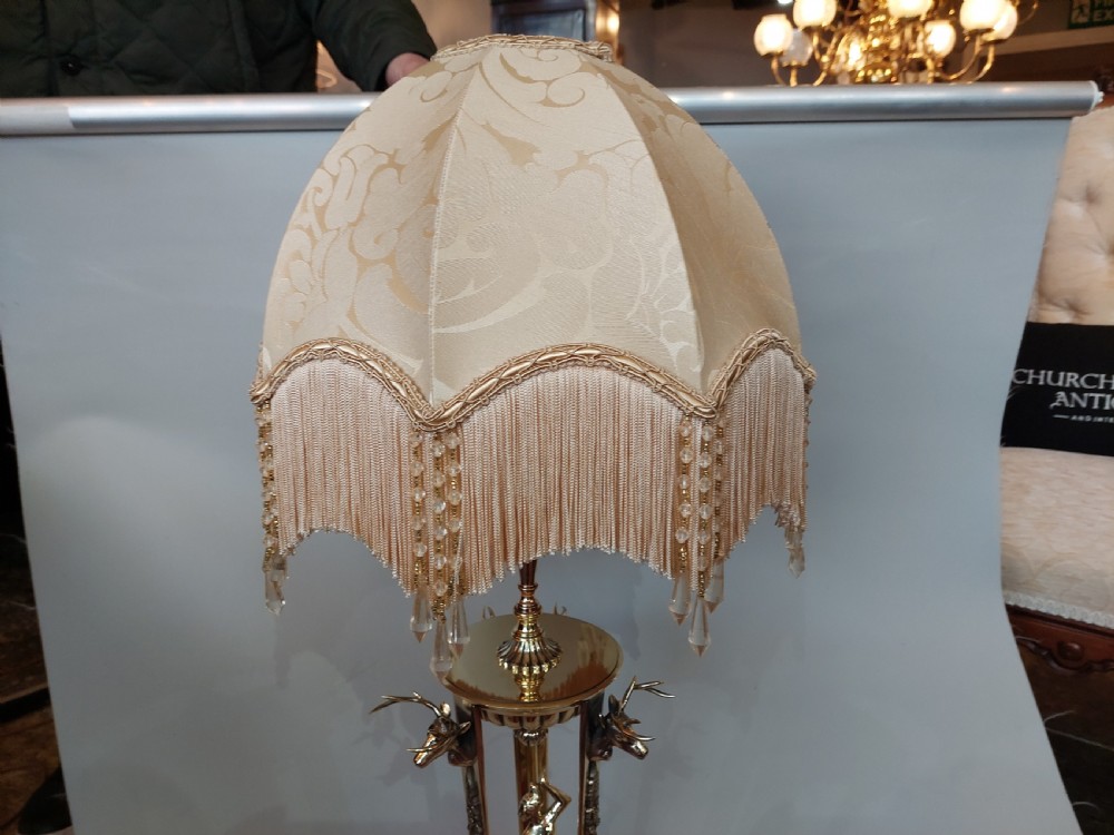An early 20th-century brass table lamp featuring a central figure of Minerva flanked by stag head mounts, topped with a cream fringed lampshade. View VII