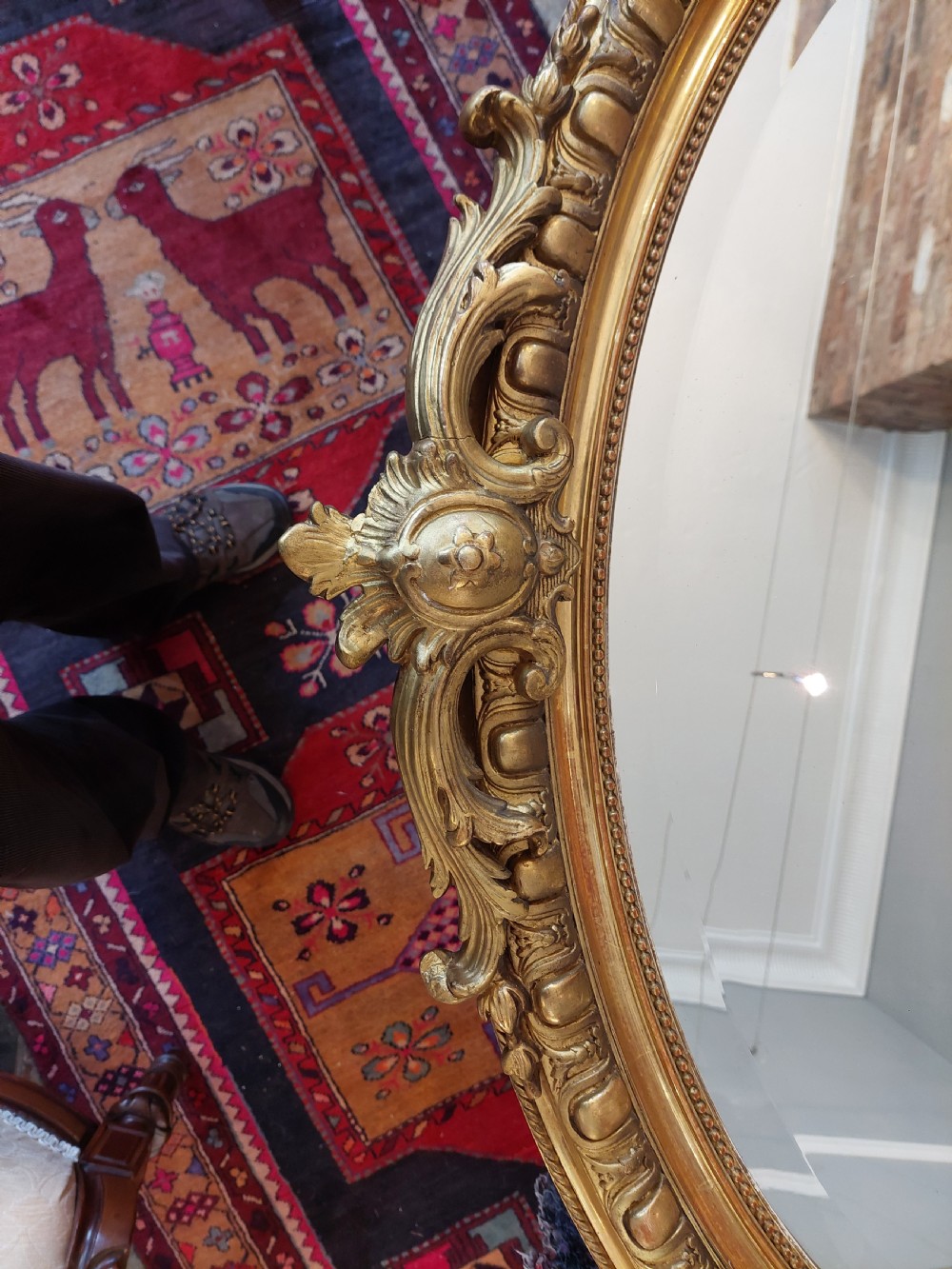 An oval Victorian Rococo style mirror featuring an ornate gilded frame with carved scrollwork, cresting, and egg-and-dart moulding. View II