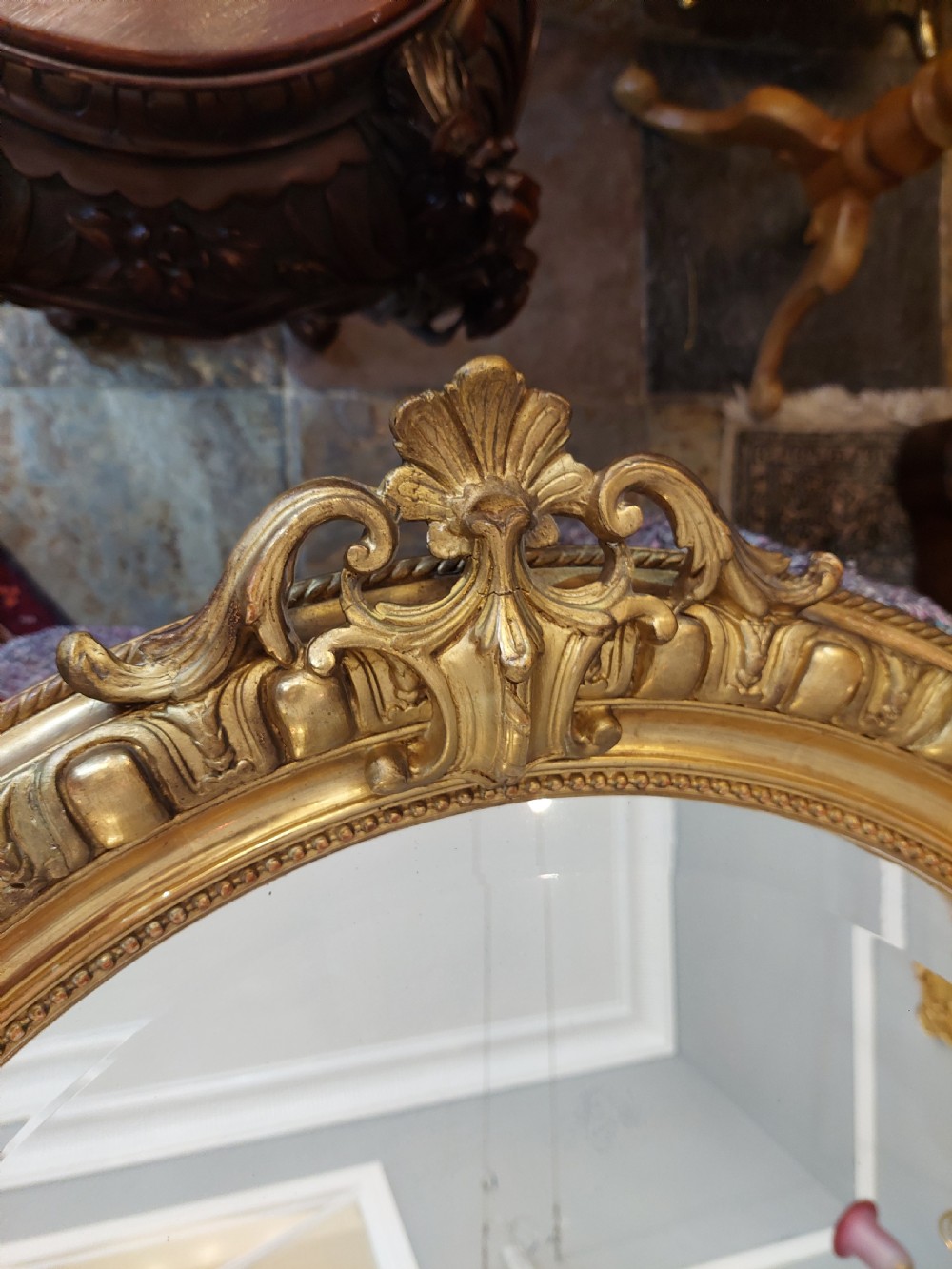 An oval Victorian Rococo style mirror featuring an ornate gilded frame with carved scrollwork, cresting, and egg-and-dart moulding. View V