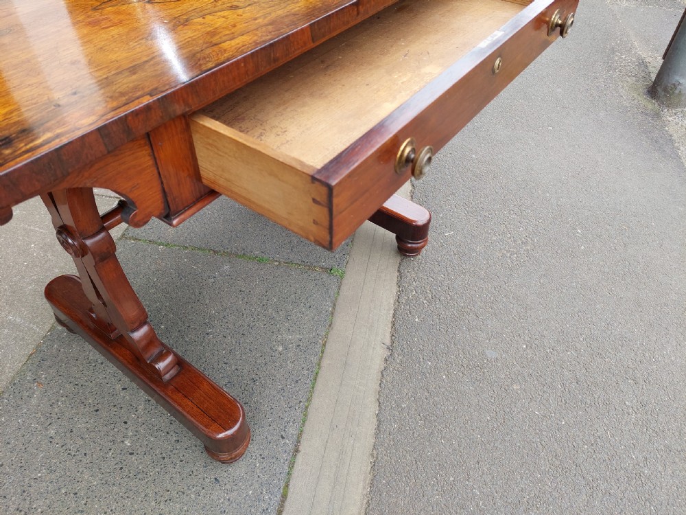 A Regency rosewood sofa table featuring a figured timber top, single frieze drawer with brass knobs, a turned central stretcher, and trestle legs. View IV