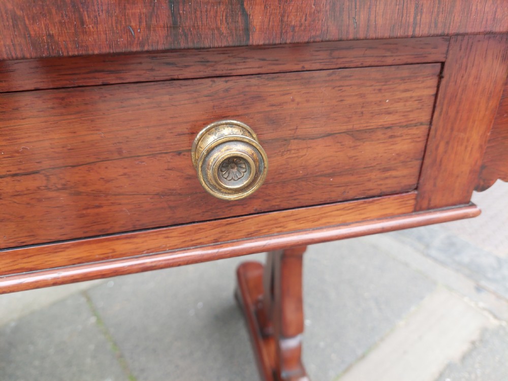 A Regency rosewood sofa table featuring a figured timber top, single frieze drawer with brass knobs, a turned central stretcher, and trestle legs. View X