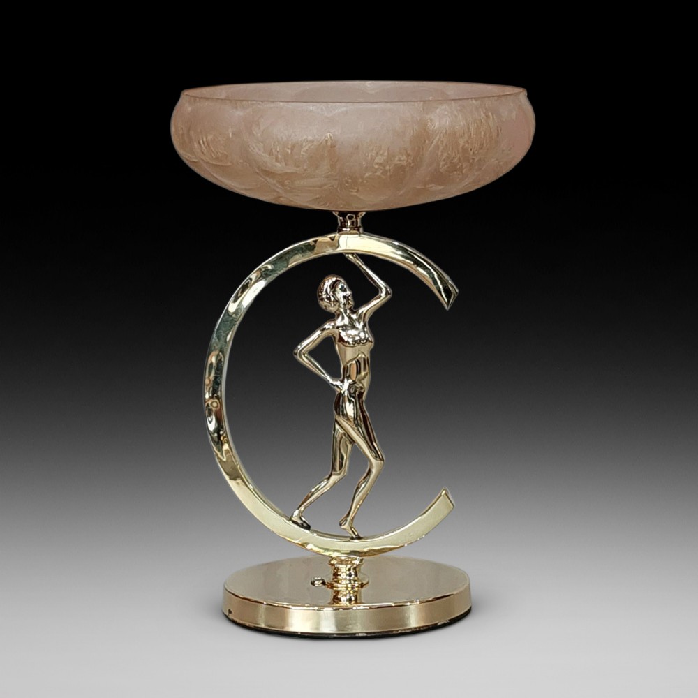 An Art Deco polished brass table lamp featuring a stylized female figure within a crescent frame supporting a pink frosted glass bowl shade.