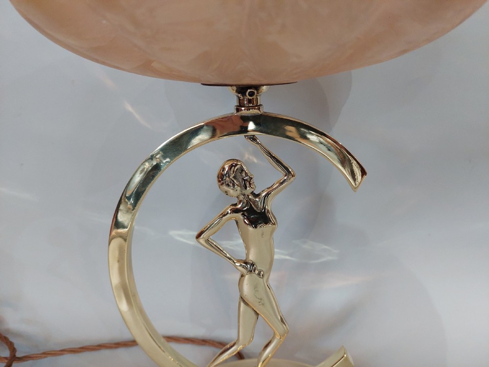 An Art Deco polished brass table lamp featuring a stylized female figure within a crescent frame supporting a pink frosted glass bowl shade. View II