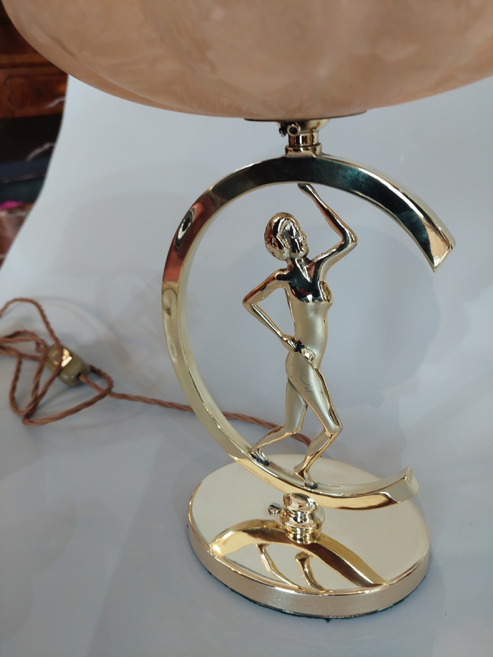 An Art Deco polished brass table lamp featuring a stylized female figure within a crescent frame supporting a pink frosted glass bowl shade. View V