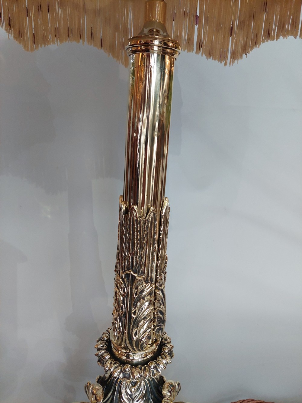 A Victorian brass table lamp featuring a columned base with acanthus leaf moulding and a cream fabric lampshade trimmed with burgundy fringe. View V