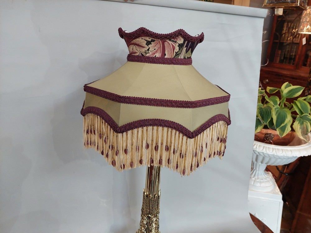 A Victorian brass table lamp featuring a columned base with acanthus leaf moulding and a cream fabric lampshade trimmed with burgundy fringe. View VII